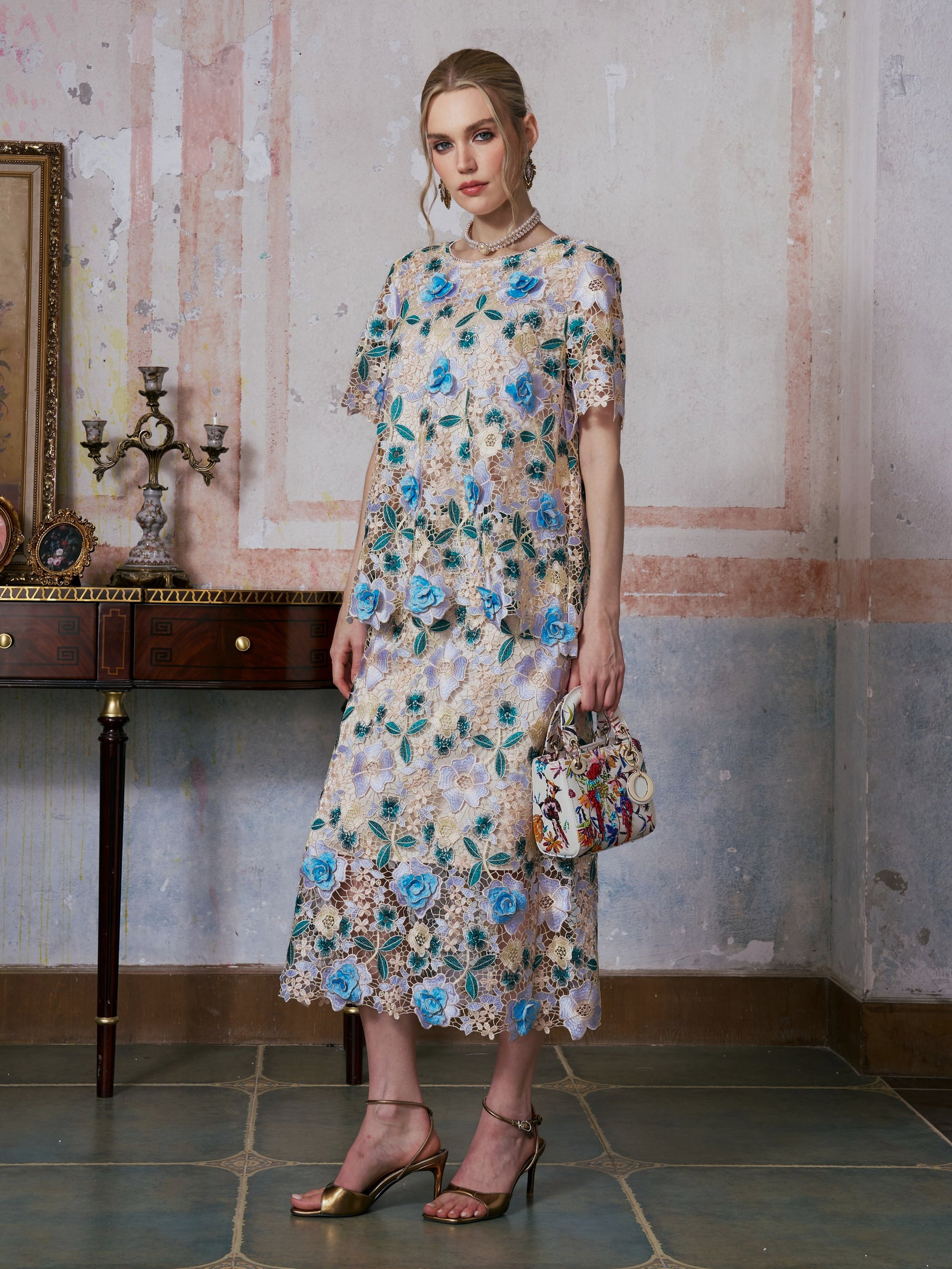 A woman stands indoors wearing the Social Queen 3D Floral Cutwork Lace Top, featuring light-toned guipure lace adorned with blue floral embroidery. She holds a small handbag and wears gold sandals, with a candelabra and framed artwork in the background.