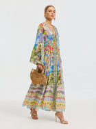 A woman in the Social Queen Garden Print Silk Maxi Dress stands against a plain light background, accessorized with a bamboo handbag and gold high-heeled sandals.