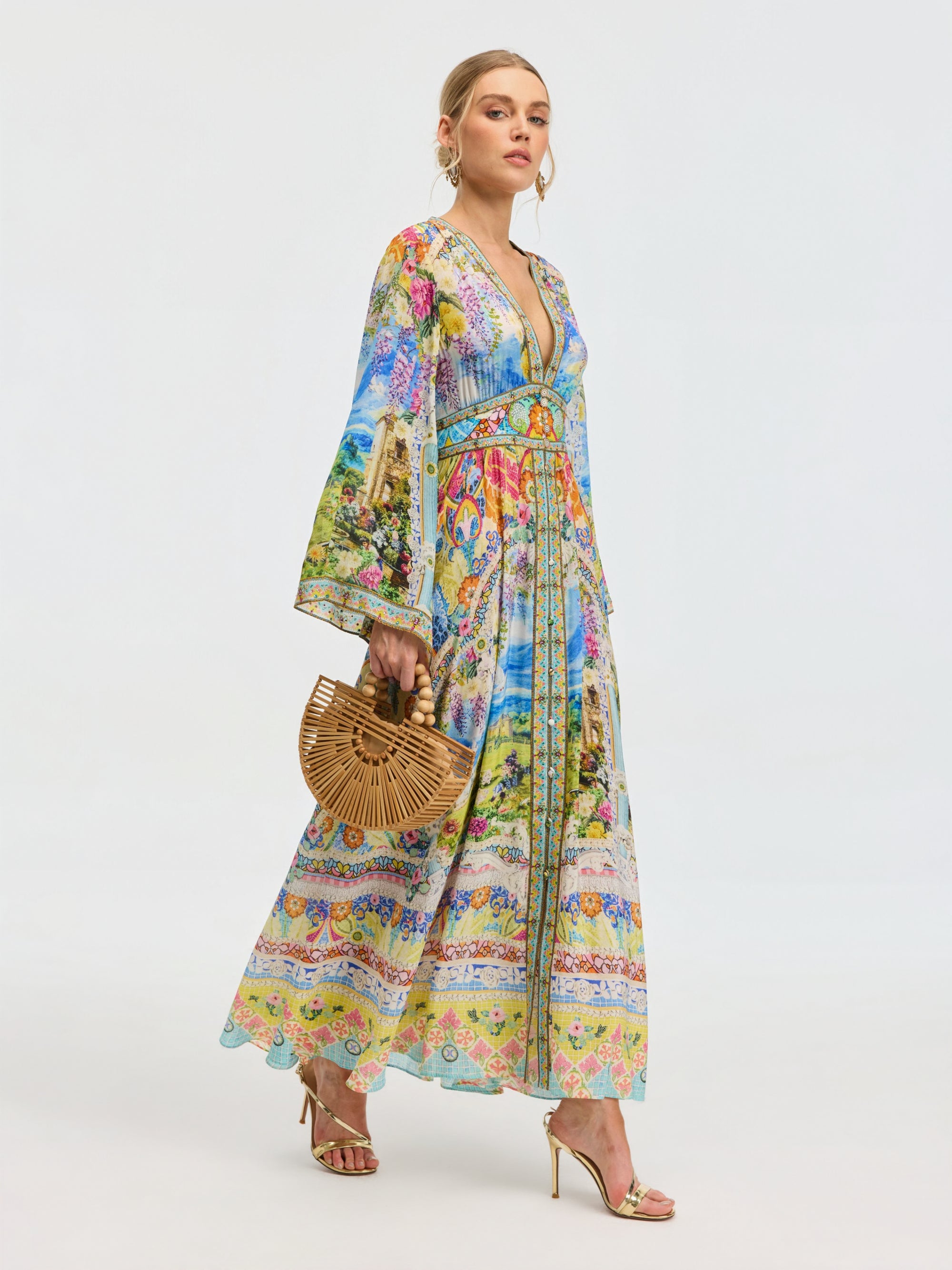 A woman in the Social Queen Garden Print Silk Maxi Dress stands against a plain light background, accessorized with a bamboo handbag and gold high-heeled sandals.