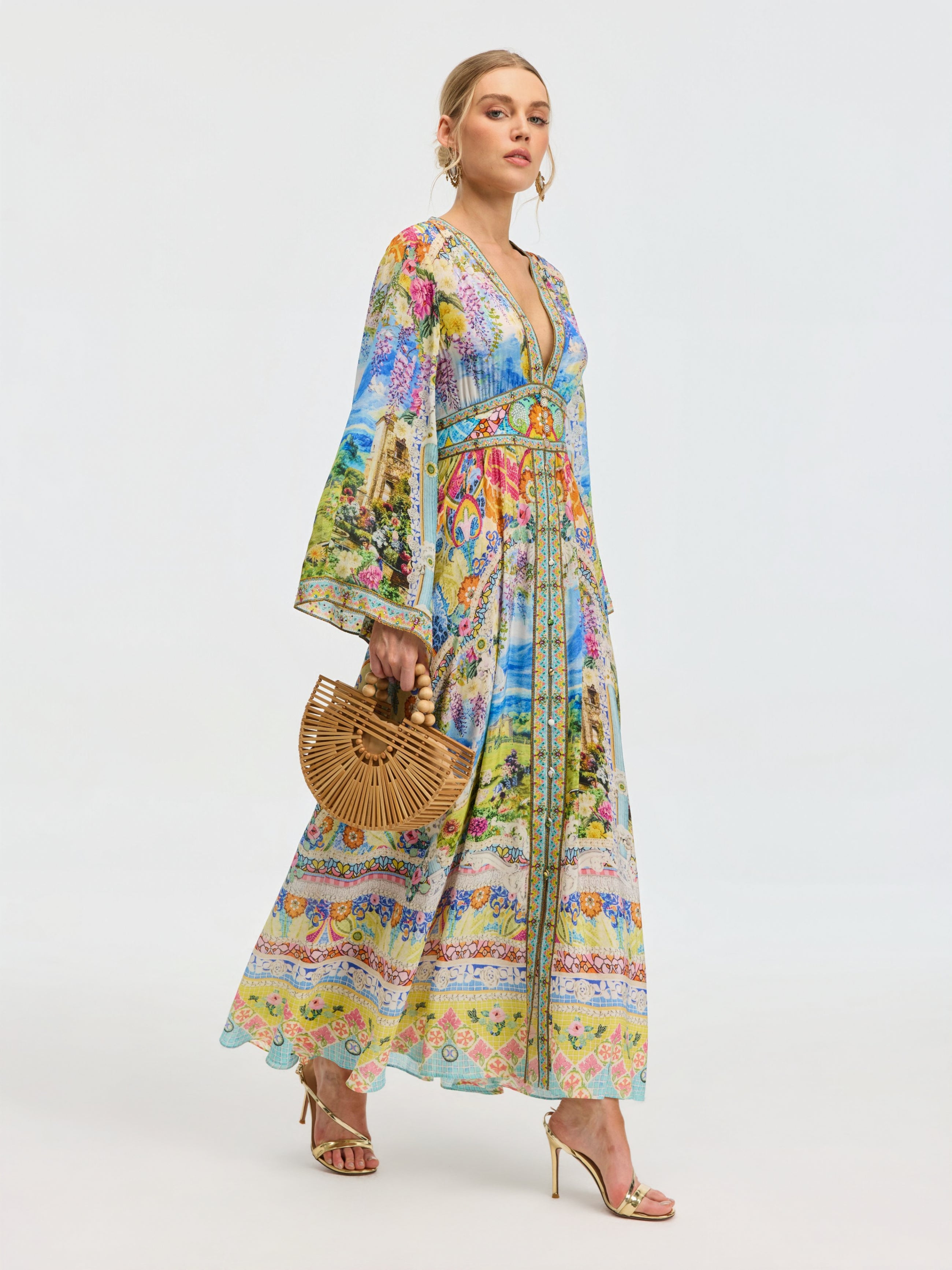 A woman in the Social Queen Garden Print Silk Maxi Dress stands against a plain light background, accessorized with a bamboo handbag and gold high-heeled sandals.