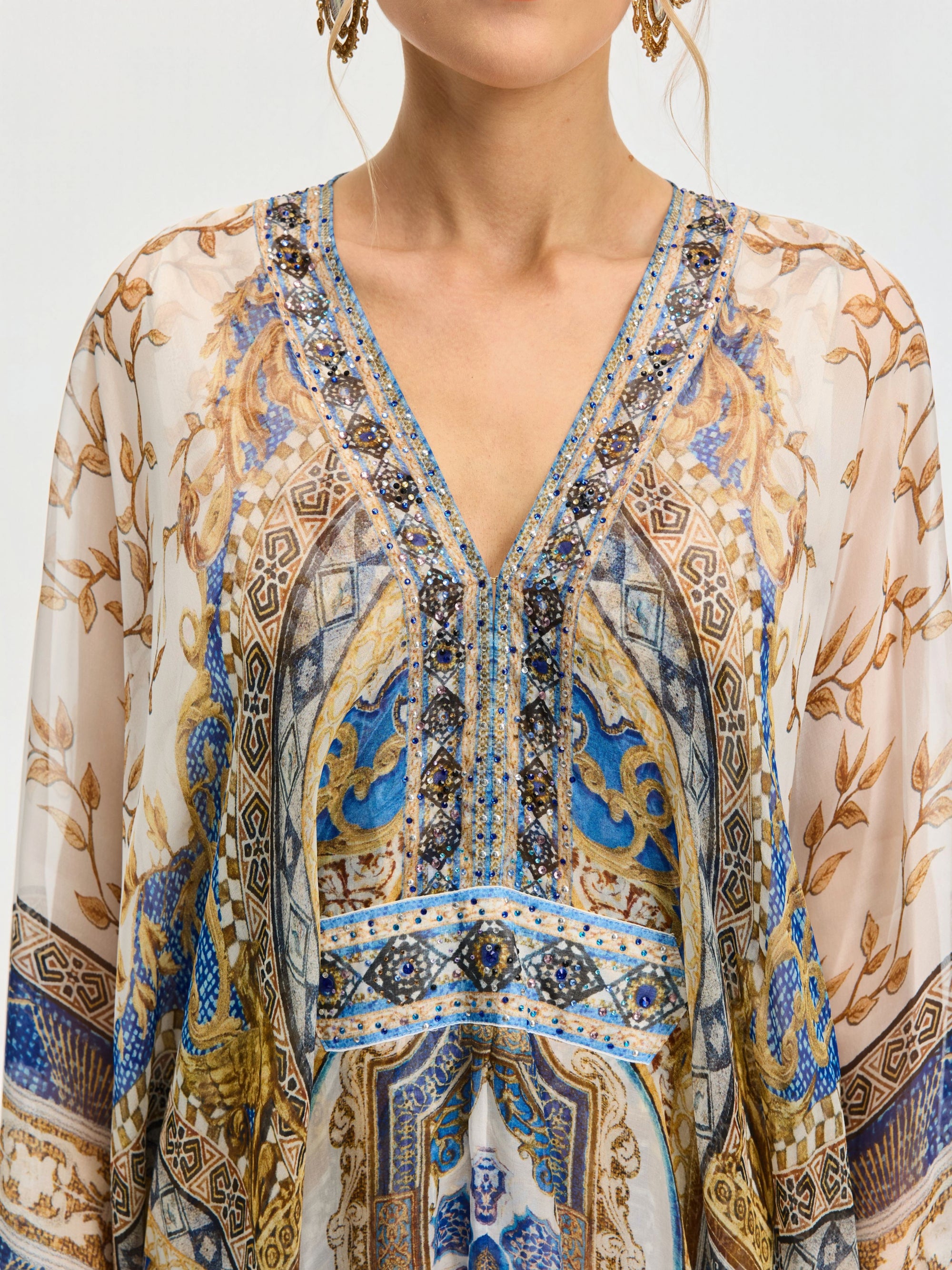 A person wears the Social Queen Byzantine Print Silk Caftan Dress, featuring a sheer blouse with blue, gold, and brown ornate patterns and a V-neckline. Only the upper torso and part of the face are visible.