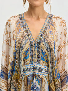 A person wears the Social Queen Byzantine Print Silk Caftan Dress, featuring a sheer blouse with blue, gold, and brown ornate patterns and a V-neckline. Only the upper torso and part of the face are visible.