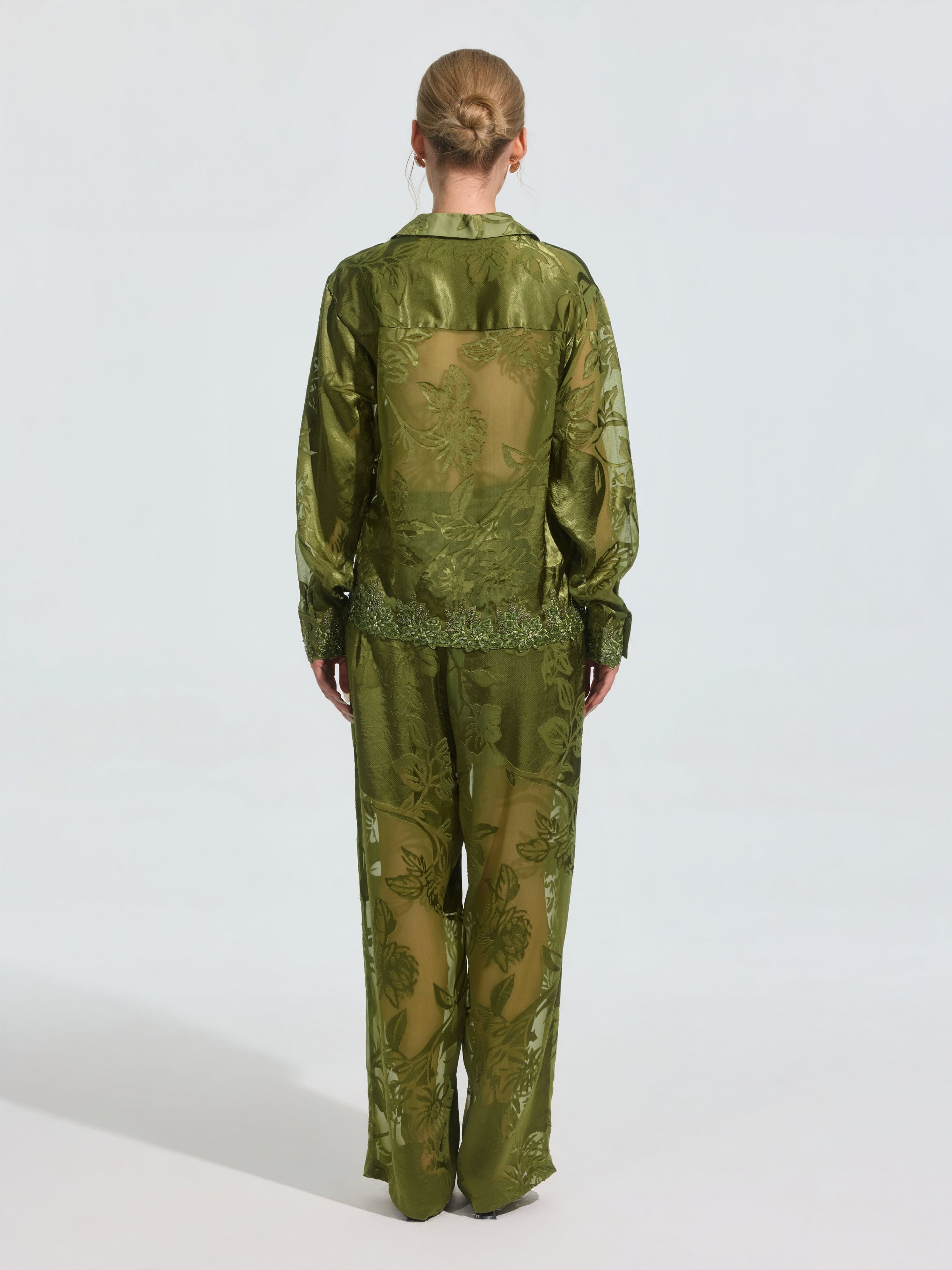 A person stands with their back turned, wearing Social Queen’s Botanical Burnout Satin Wide-Leg Pants in green with a botanical pattern, paired with a sheer floral blouse and hair tied back.