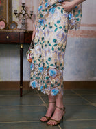A person stands indoors wearing the Social Queen 3D Floral Cutwork Lace Skirt in gold and blue, paired with gold heels and holding a small handbag. The background features a table, candleholder, and framed mirror.