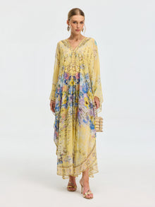 A woman stands against a white background wearing the Social Queen Floral Print Silk Caftan Dress with long sleeves, holding a beaded handbag—perfect resort wear for effortless elegance.