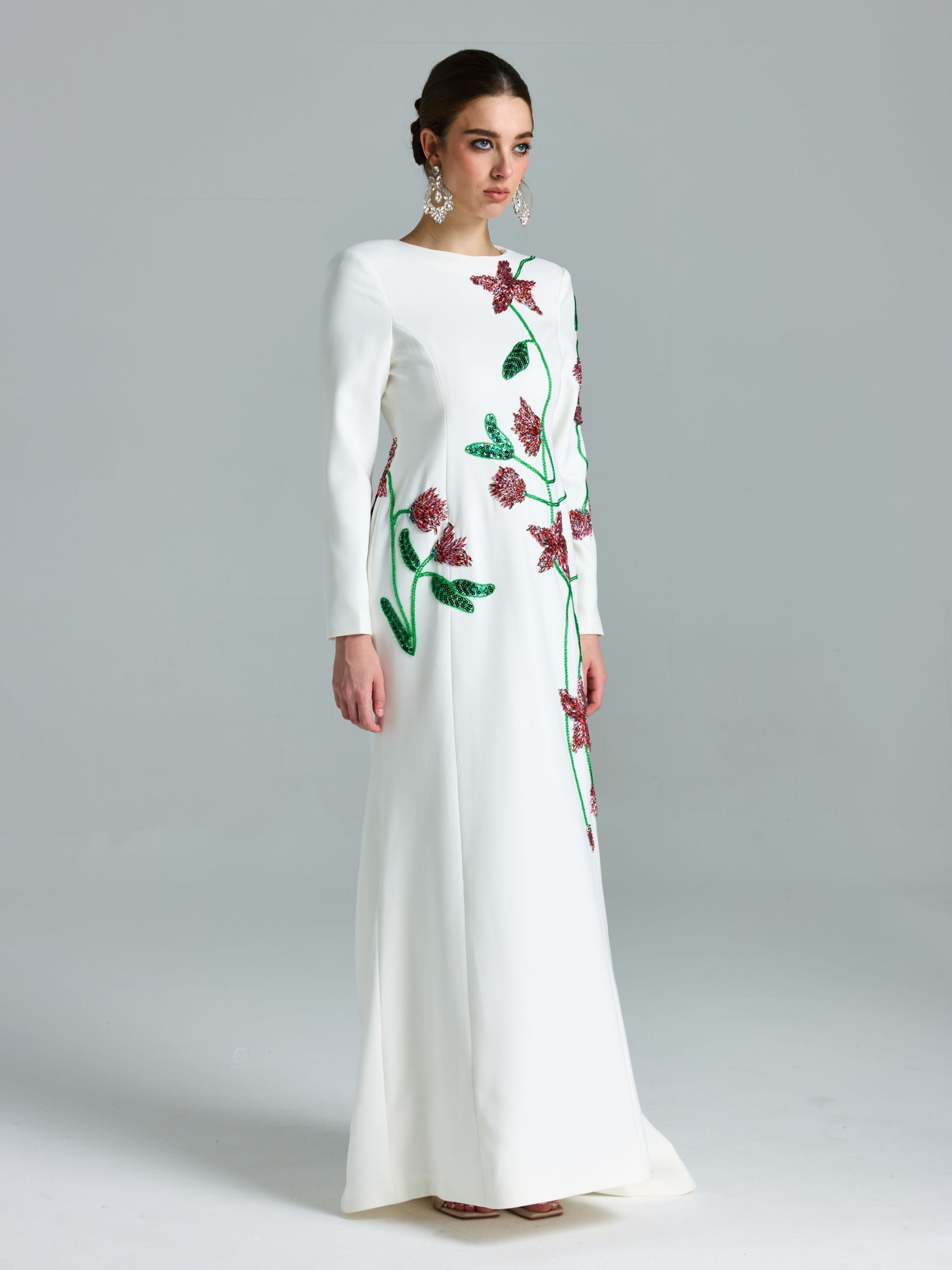 A woman wears the Social Queen Floral Beaded A-Line Gown with green stems and pink floral embroidery, featuring long sleeves and a floor-length A-line silhouette, posing against a plain gray background.