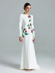 A woman wears the Social Queen Floral Beaded A-Line Gown with green stems and pink floral embroidery, featuring long sleeves and a floor-length A-line silhouette, posing against a plain gray background.