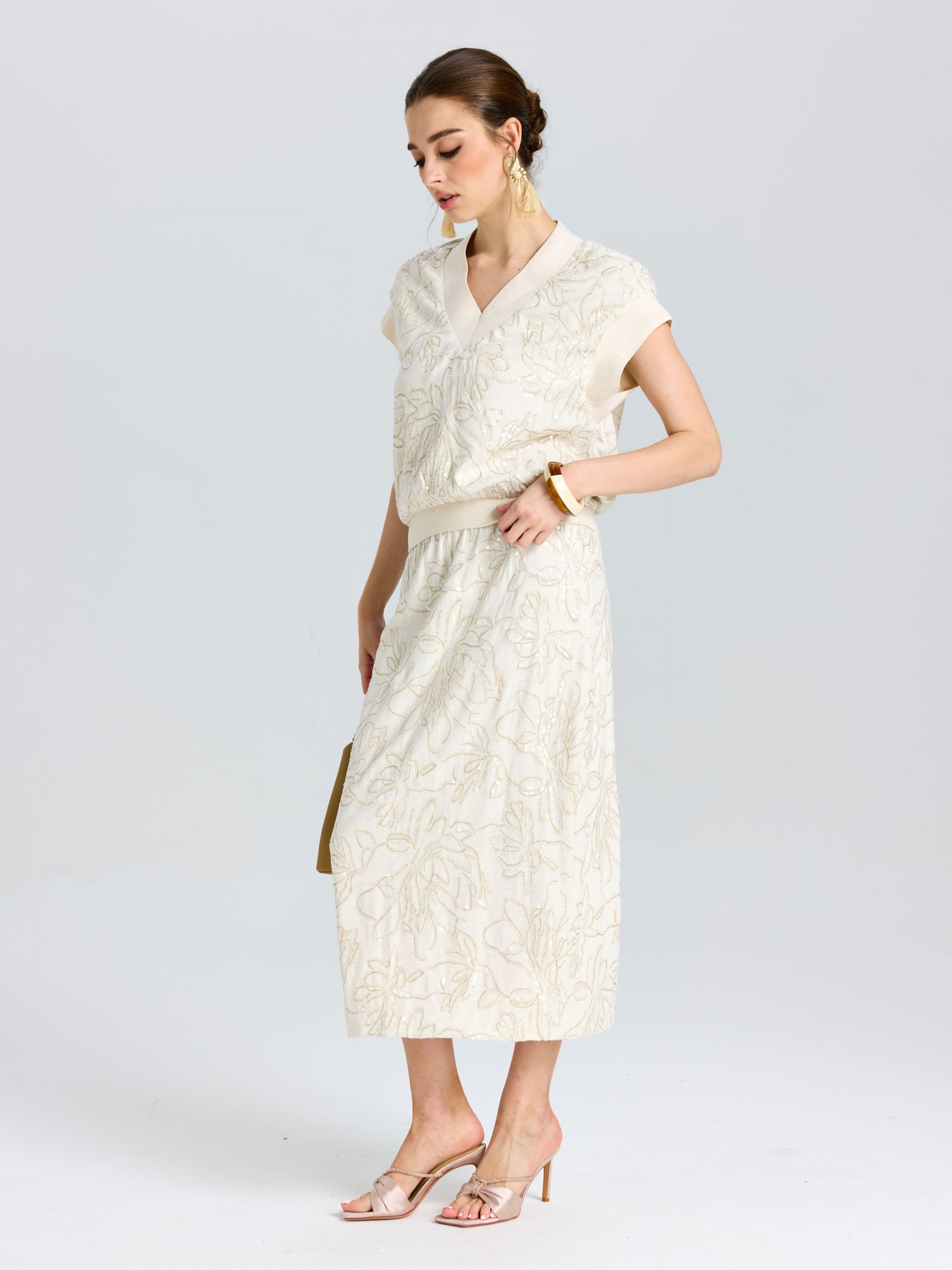 A woman stands in profile wearing the Social Queen Floral Embroidered Linen Midi Skirt with a cream short-sleeve blouse, beige heels, gold earrings, and a bracelet.