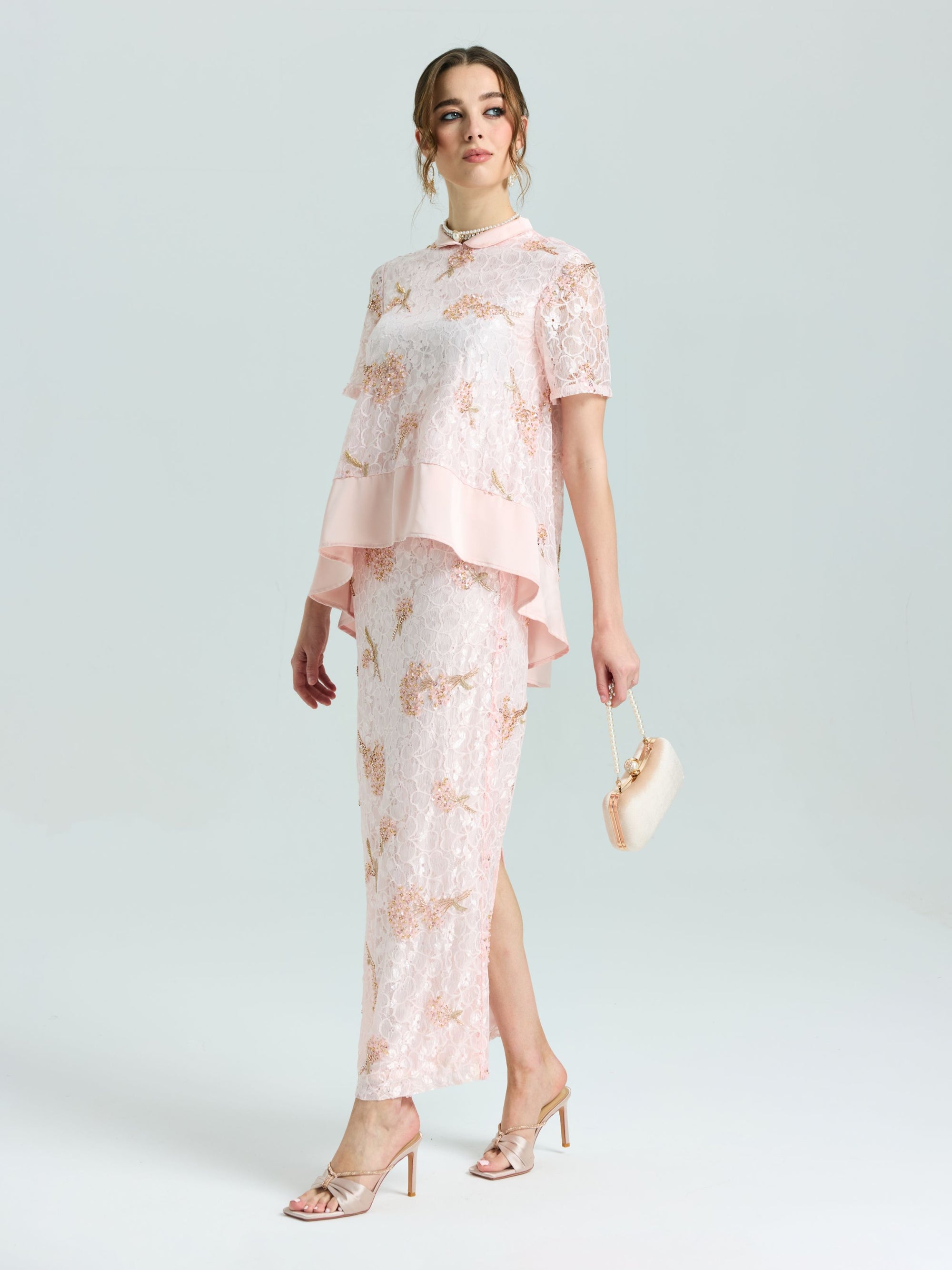 A woman wears the Social Queen Dragonfly Beaded Lace Midi Skirt in pale pink with a straight silhouette, styled with a small beige purse and heeled sandals, standing against a plain light background.