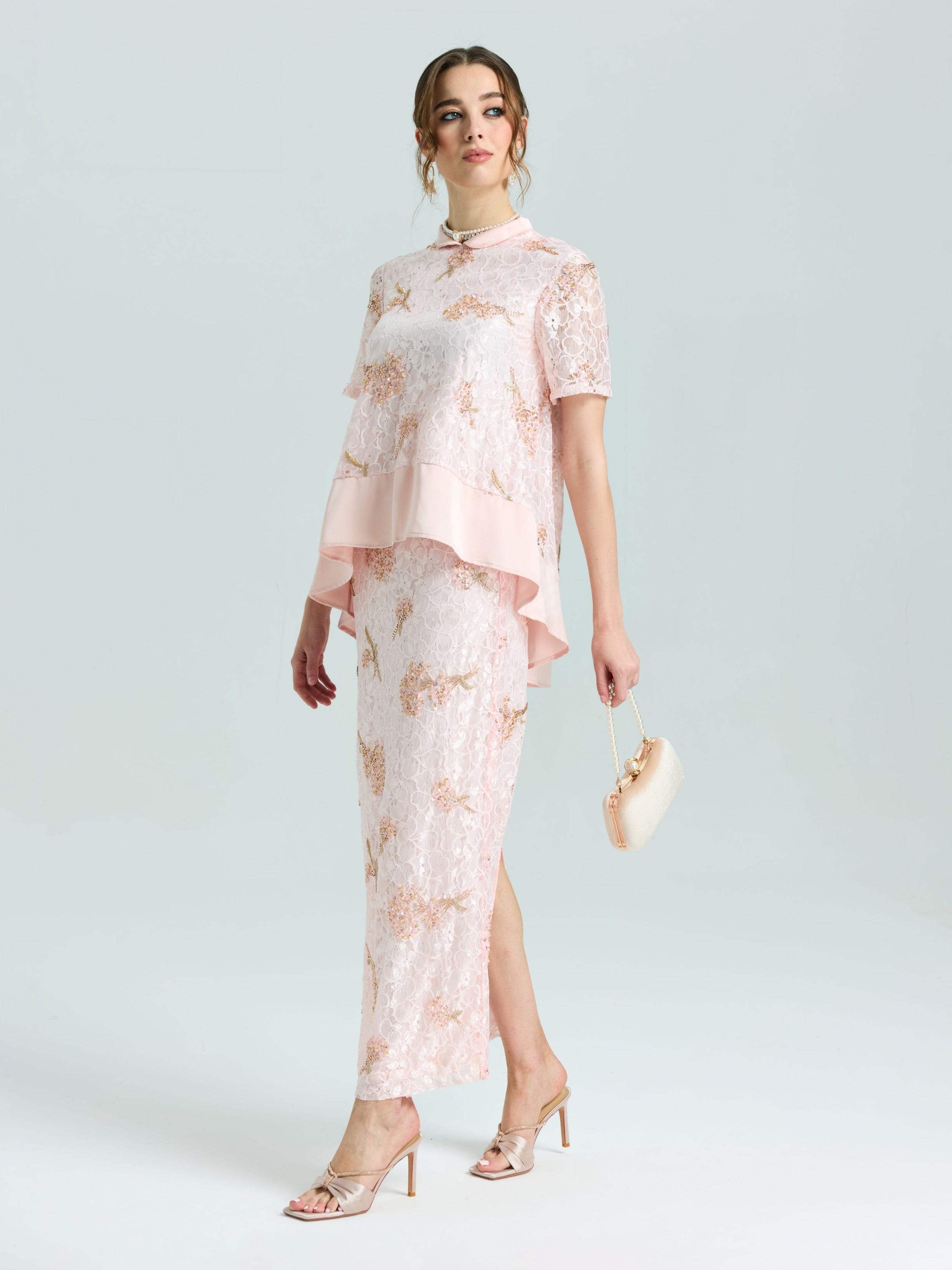 A woman wears the Social Queen Dragonfly Beaded Lace Midi Skirt in pale pink with a straight silhouette, styled with a small beige purse and heeled sandals, standing against a plain light background.