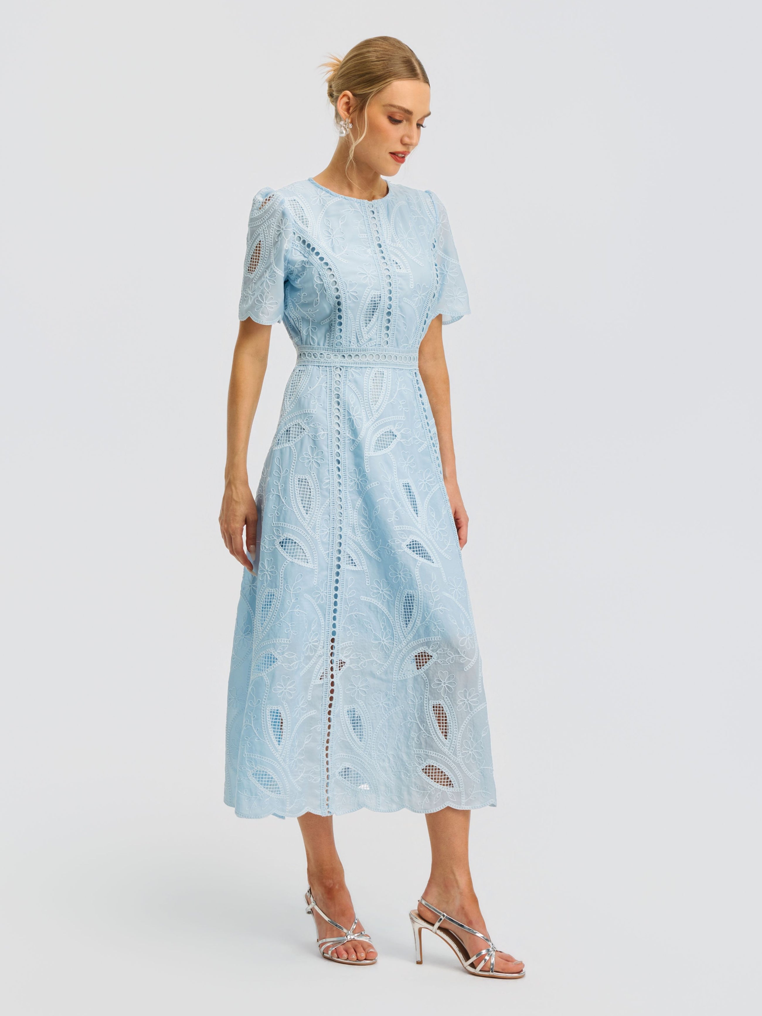 A woman wearing the Social Queen Floral Cutwork Embroidered Dress—featuring a light blue lace midi silhouette with short sleeves and delicate floral embroidery—is paired with silver heeled sandals, standing against a plain white background.