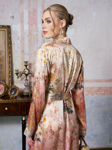 A woman wearing the Social Queen Garden Print Mulberry Silk Shirt Dress stands in a vintage-style room, glancing over her shoulder; the baroque garden print of her dress mirrors the charm of antique furniture and framed art in the background.