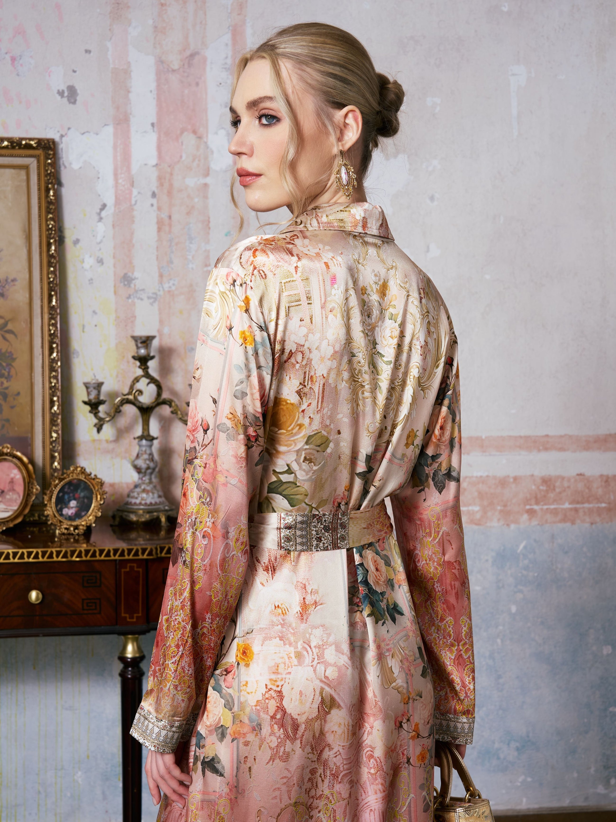 A woman wearing the Social Queen Garden Print Mulberry Silk Shirt Dress stands in a vintage-style room, glancing over her shoulder; the baroque garden print of her dress mirrors the charm of antique furniture and framed art in the background.