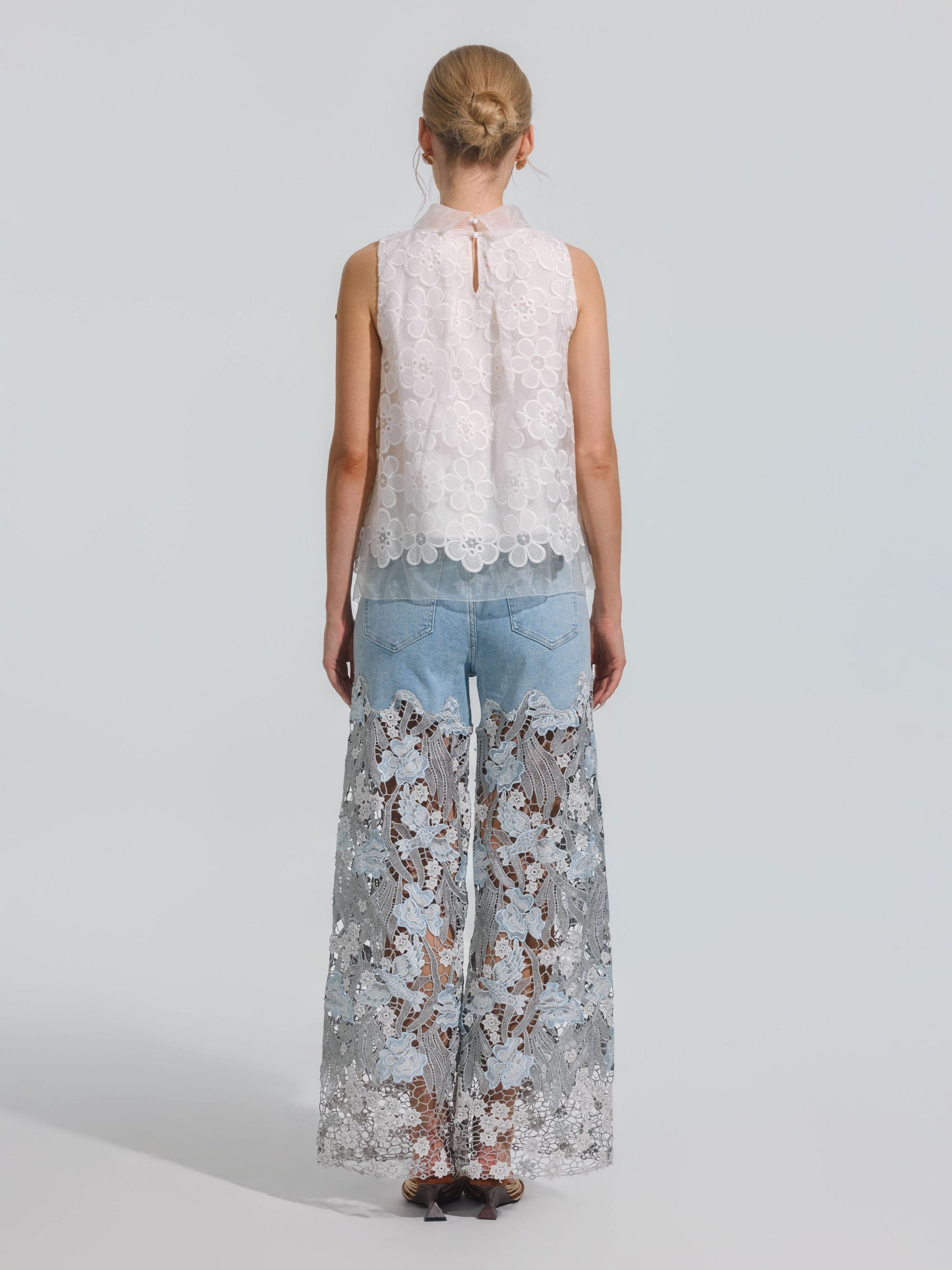 A woman is seen from behind, wearing the Social Queen Floral Lace Patchwork Pants—a sheer, wide-leg style featuring a white lace overlay and blue stretch denim shorts underneath—paired with a sleeveless white lace top, against a plain light background.