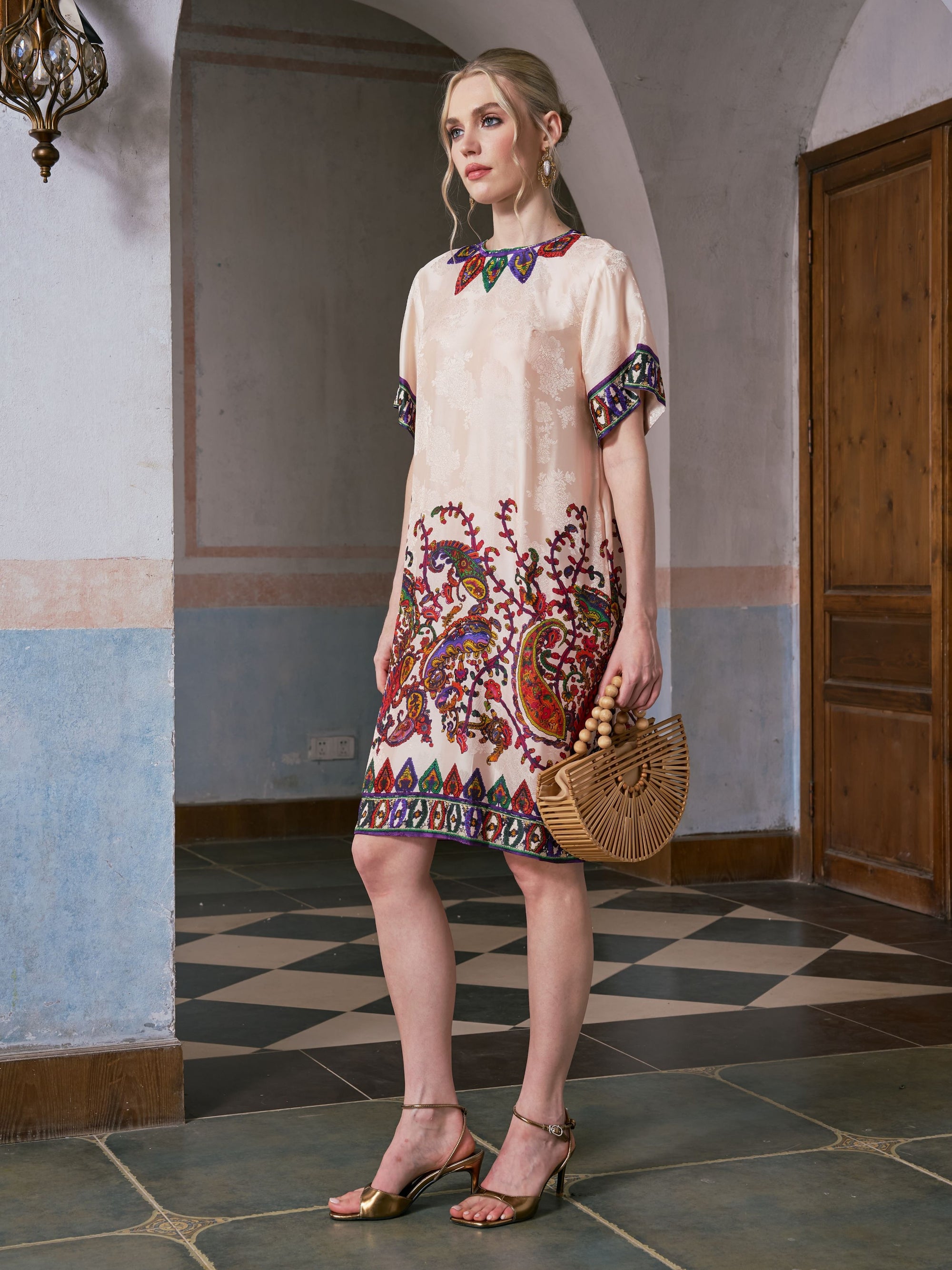 A woman wearing the Social Queen Paisley Garden Print Jacquard Silk Dress and gold heels holds a bamboo handbag, standing indoors on a tiled floor near a wooden door.