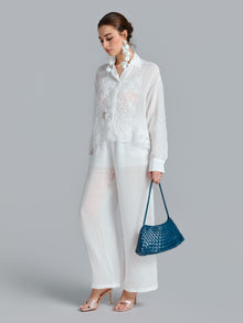 A woman stands against a plain grey background, wearing the Social Queen 3D Lace Appliqué Shirt styled with matching pants, large white earrings, nude heels, and holding a blue woven handbag.