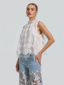 A woman models the Social Queen 3D Floral Lace Appliqué Silk Top—an elegant sleeveless white piece adorned with delicate 3D floral details—paired with light blue jeans featuring sheer lace floral panels, set against a plain light gray background.