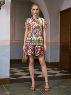 A woman stands indoors wearing Social Queen's Paisley Garden Print Silk Shorts, paired with a wicker handbag and heeled sandals. She poses against a tiled floor and plain, arched wall background, embodying effortless resort wear style.