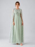A woman wearing the Social Queen Beaded Printed Cape Chiffon Dress—an elegant, long maxi occasion dress in light green chiffon with floral embellishments and a sheer, beaded cape—stands against a plain grey background.