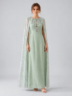 A woman wearing the Social Queen Beaded Printed Cape Chiffon Dress—an elegant, long maxi occasion dress in light green chiffon with floral embellishments and a sheer, beaded cape—stands against a plain grey background.