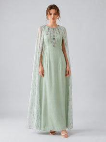 A woman wearing the Social Queen Beaded Printed Cape Chiffon Dress—an elegant, long maxi occasion dress in light green chiffon with floral embellishments and a sheer, beaded cape—stands against a plain grey background.