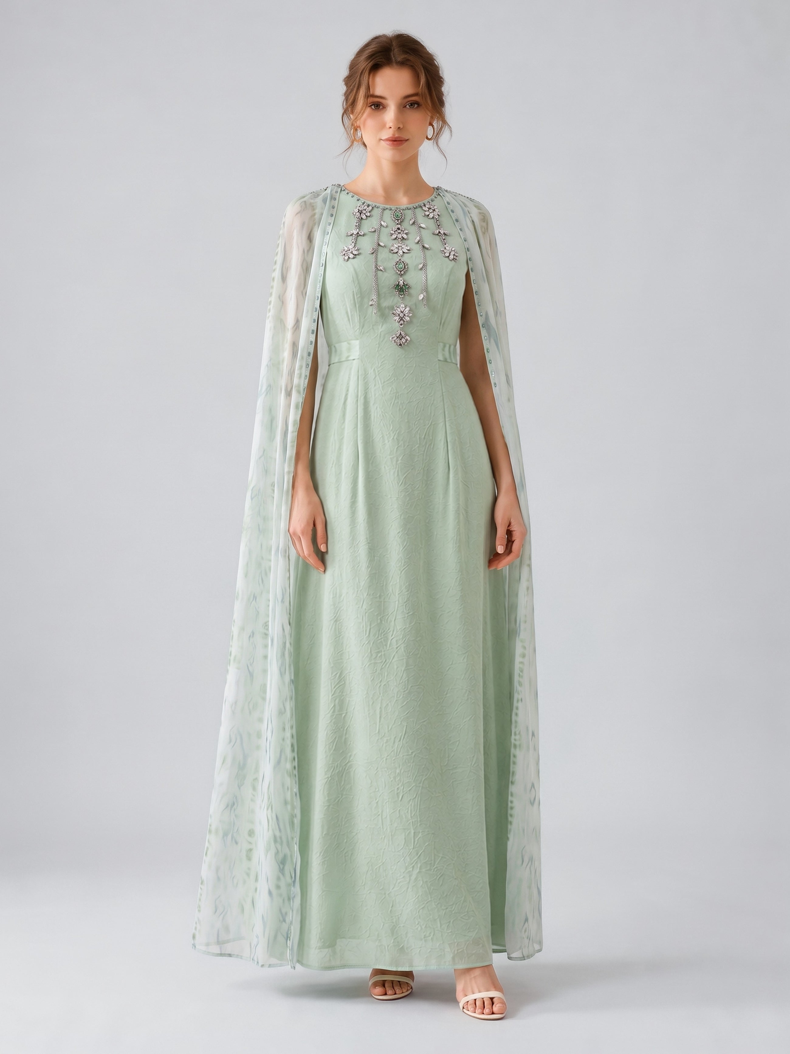 A woman wearing the Social Queen Beaded Printed Cape Chiffon Dress—an elegant, long maxi occasion dress in light green chiffon with floral embellishments and a sheer, beaded cape—stands against a plain grey background.