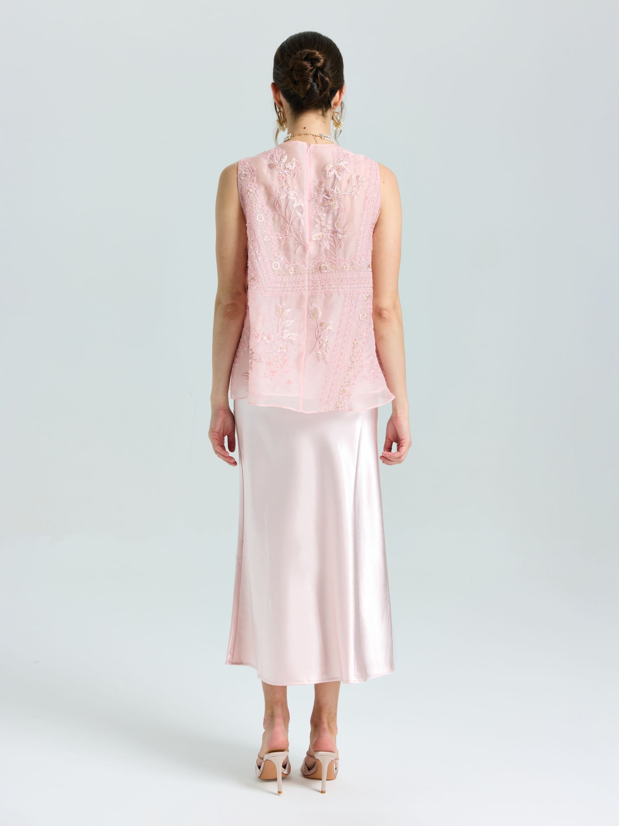 A woman faces away, wearing a light pink sleeveless lace top and the Satin A-Line Midi Skirt in pale pink by Social Queen, paired with heeled sandals.