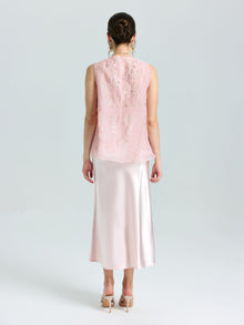 A woman faces away, wearing a light pink sleeveless lace top and the Satin A-Line Midi Skirt in pale pink by Social Queen, paired with heeled sandals.