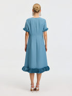 A woman stands facing away, wearing the Social Queen Beaded Crystal Embellished Dress, a light blue, knee-length design with ruffled sleeves and hem, beaded neckline details, paired with white high-heeled sandals.