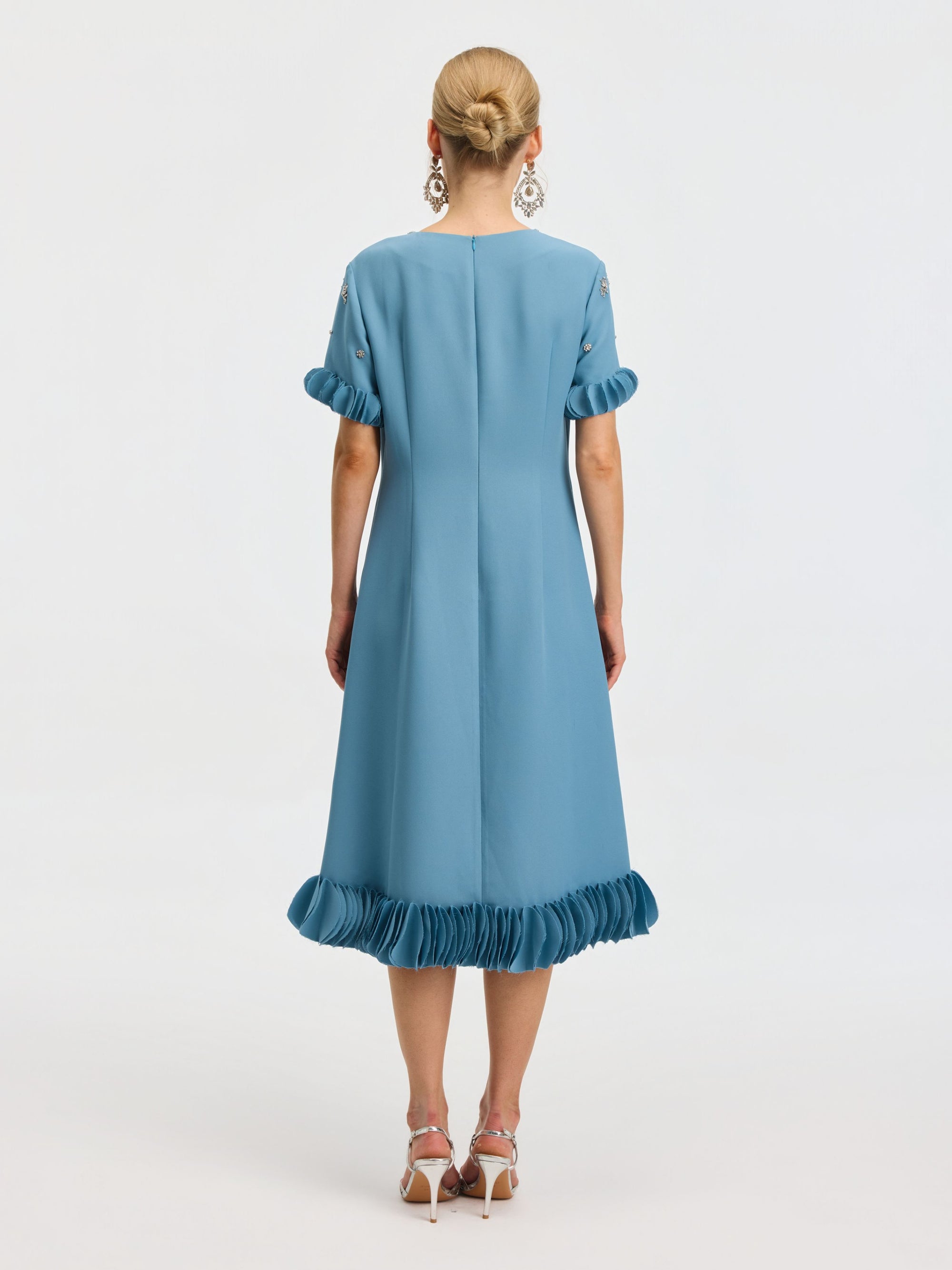 A woman stands facing away, wearing the Social Queen Beaded Crystal Embellished Dress, a light blue, knee-length design with ruffled sleeves and hem, beaded neckline details, paired with white high-heeled sandals.
