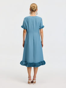 A woman stands facing away, wearing the Social Queen Beaded Crystal Embellished Dress, a light blue, knee-length design with ruffled sleeves and hem, beaded neckline details, paired with white high-heeled sandals.