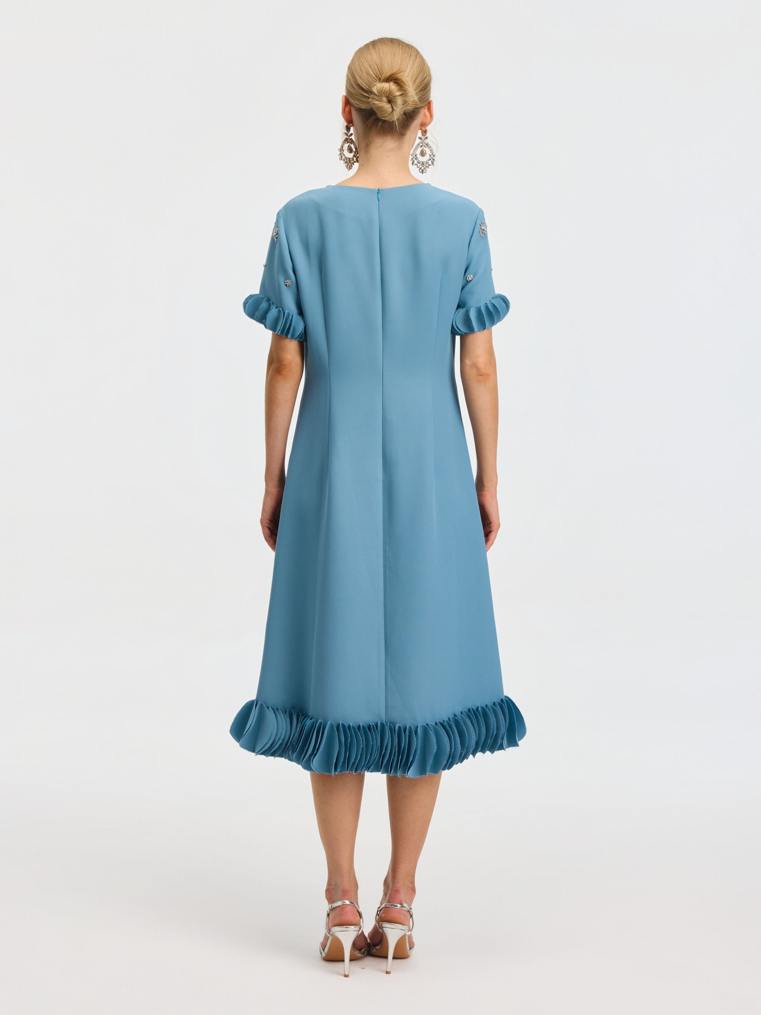 A woman stands facing away, wearing the Social Queen Beaded Crystal Embellished Dress, a light blue, knee-length design with ruffled sleeves and hem, beaded neckline details, paired with white high-heeled sandals.