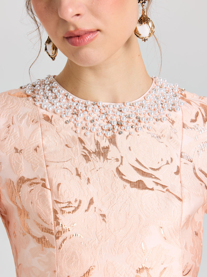 A woman wears the Social Queen Pearl Embellished Jacquard Dress in light pink with a metallic rose motif and pearl neckline, styled with gold hoop earrings and wavy hair.