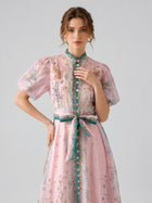 A woman stands against a plain background wearing the Social Queen Peacock Botanical Print Dress, featuring a light pink floral pattern, A-line silhouette, puff sleeves, green button placket, and a matching belt.