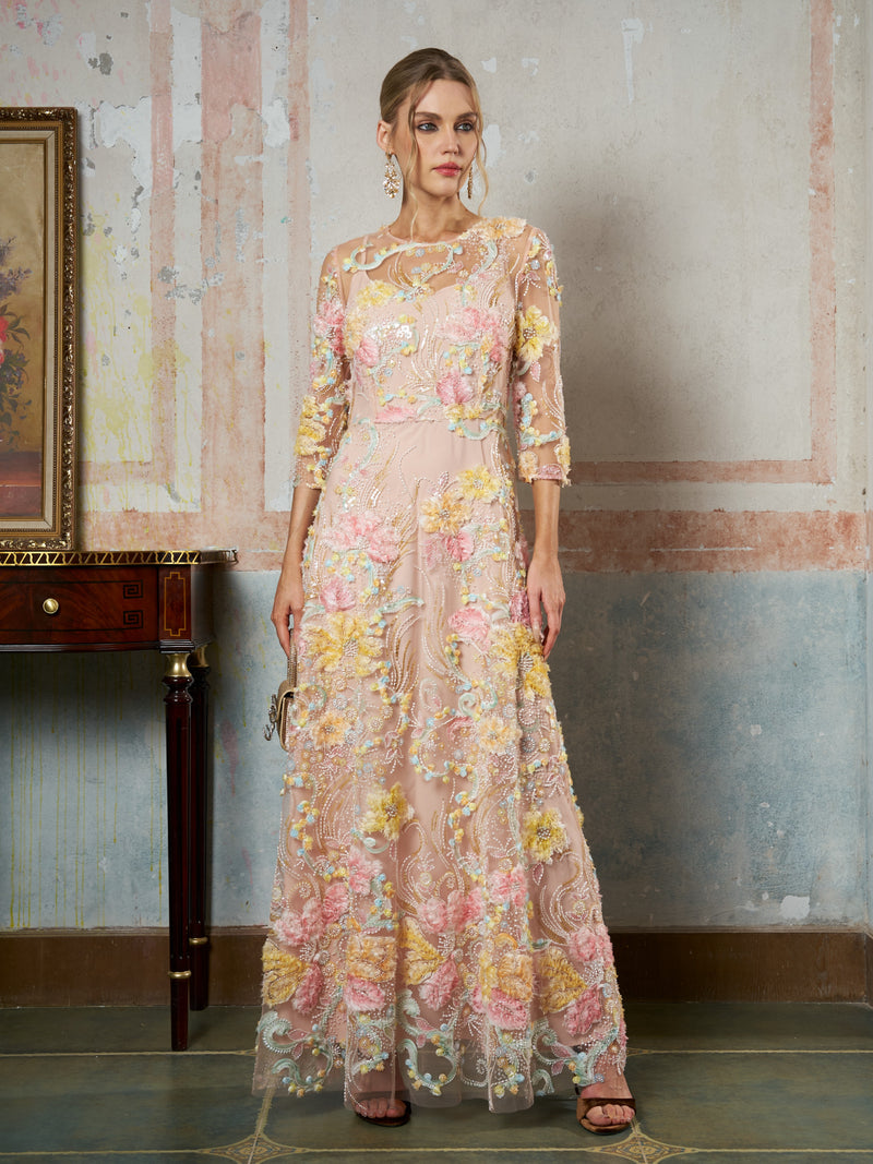A woman indoors wears the Social Queen Floral Beaded Sheer Gown, a long pastel evening dress with 3D floral appliqué and sheer sleeves, holds a clutch, and looks at the camera.