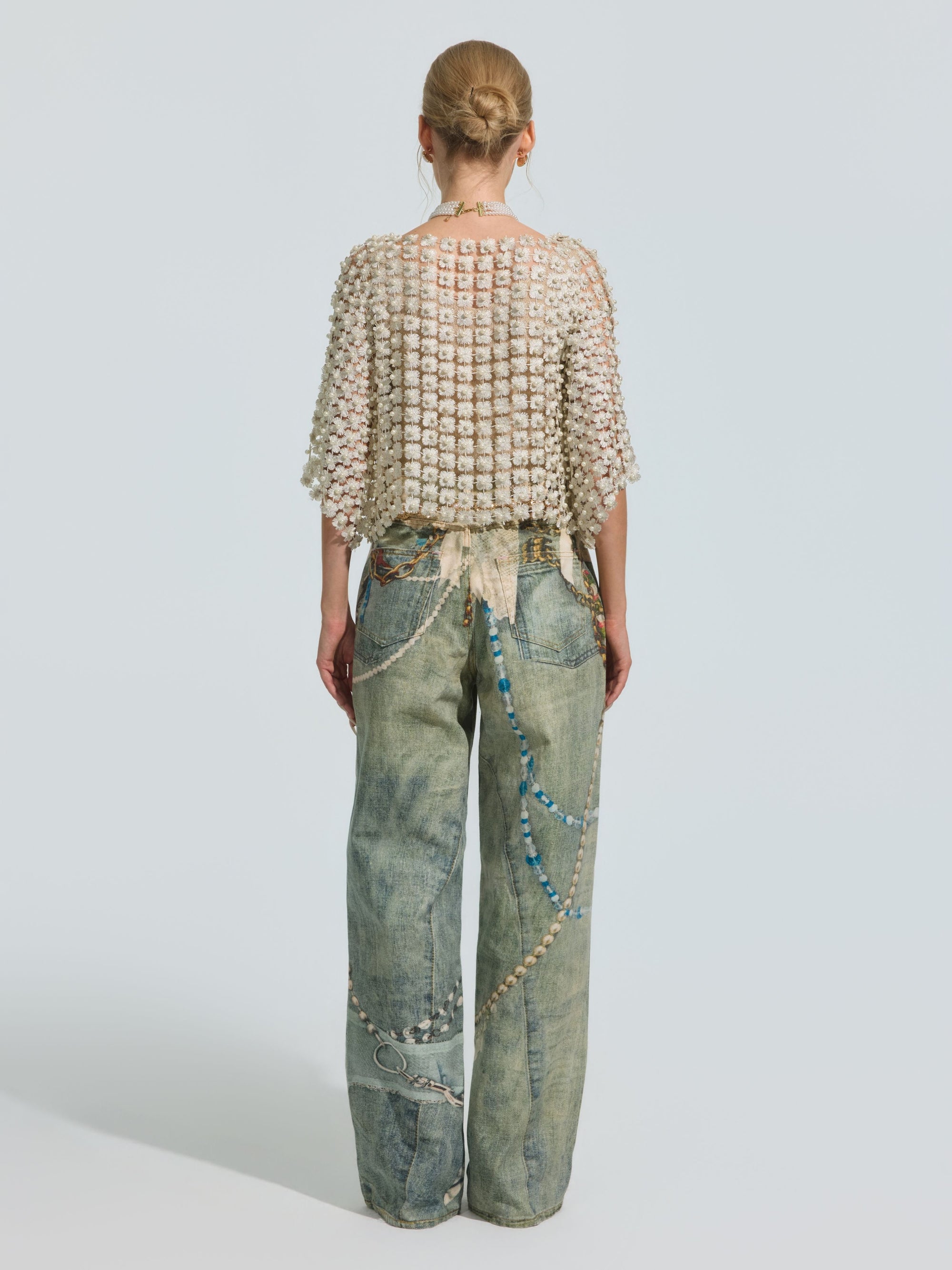 A person stands with their back to the camera, wearing Social Queen’s Pearl Chain Printed Jeans in a wide-leg silhouette, paired with a textured cream top and finished with beaded accessories draped from the waist to the pocket, set against a plain light background.