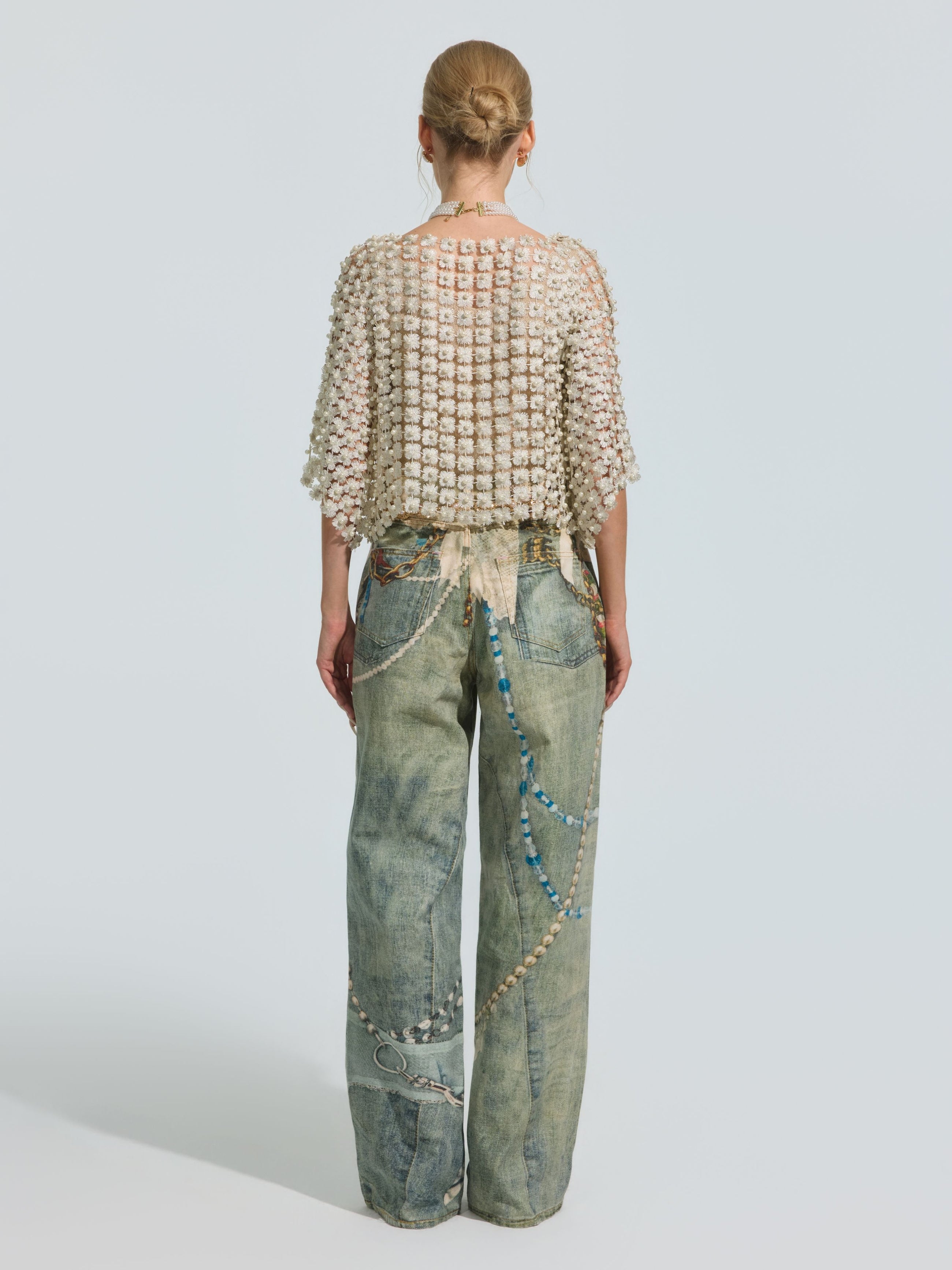 A person stands with their back to the camera, wearing Social Queen’s Pearl Chain Printed Jeans in a wide-leg silhouette, paired with a textured cream top and finished with beaded accessories draped from the waist to the pocket, set against a plain light background.