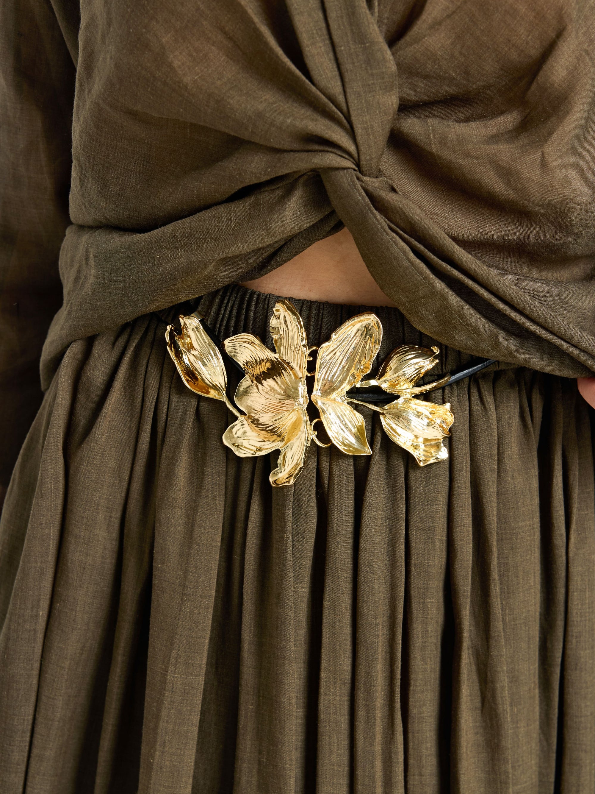 Close-up of a person in an olive-brown outfit with a twisted top and pleated skirt, styled with Social Queen's Gilded Magnolia Petal Belt in black and gold at the waist.