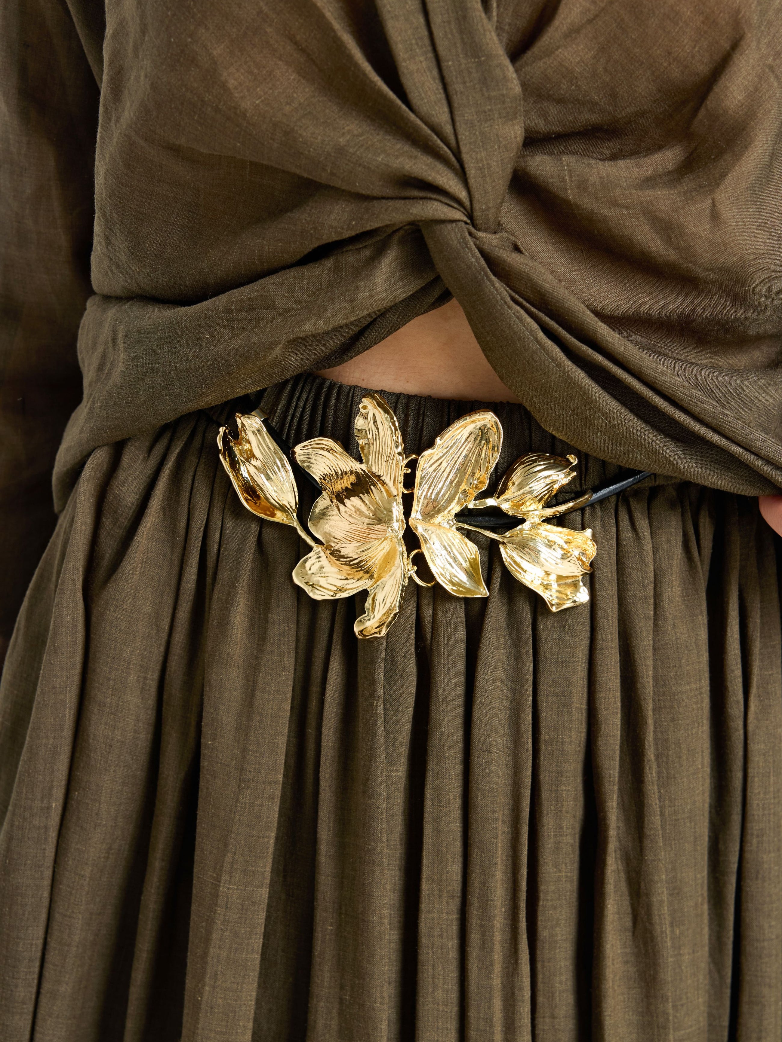 Close-up of a person in an olive-brown outfit with a twisted top and pleated skirt, styled with Social Queen's Gilded Magnolia Petal Belt in black and gold at the waist.
