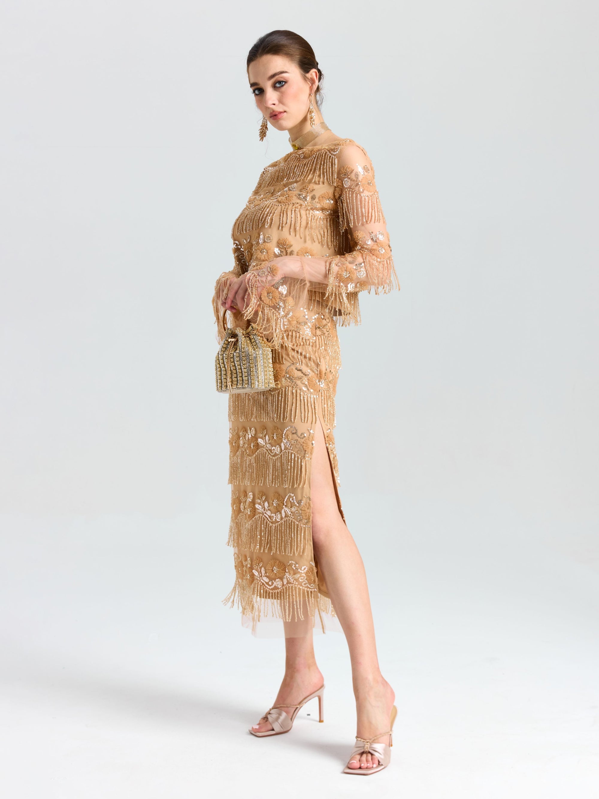 A woman wears the Social Queen Beaded Fringe Slit Midi Skirt and heels, holding a small woven handbag, standing against a plain light background.