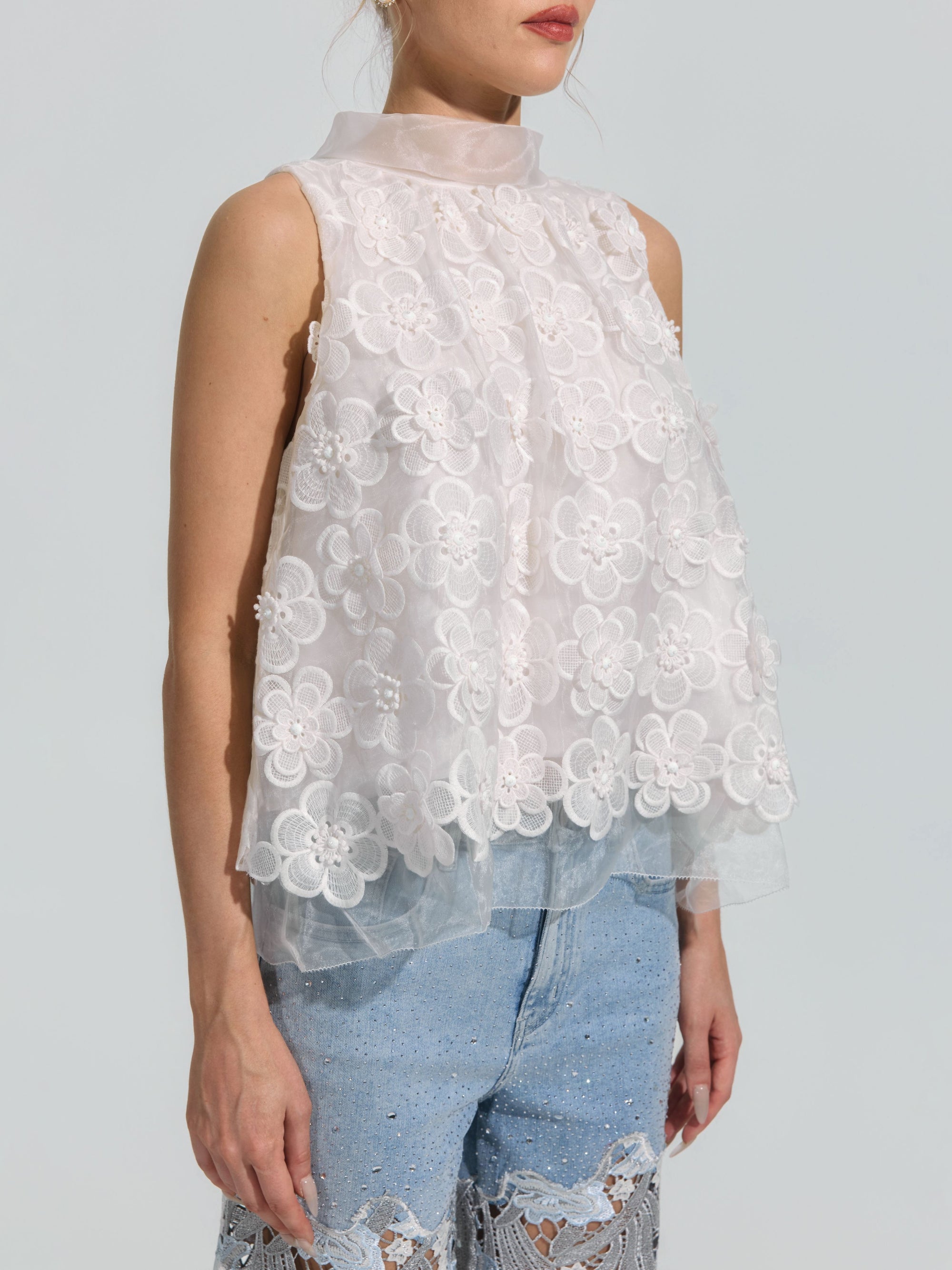 A woman wearing the Social Queen 3D Floral Lace Appliqué Silk Top, a sleeveless white blouse with a high collar and delicate lace appliqué throughout, paired with light blue jeans embellished with intricate lace and sequin details.
