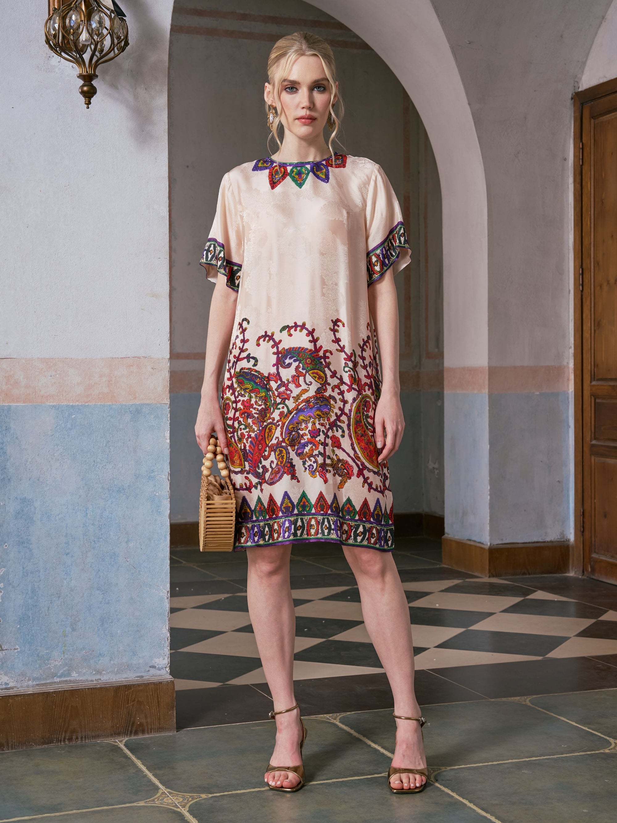 A woman wearing the Social Queen Paisley Garden Print Jacquard Silk Dress stands on a tiled floor in an arched indoor setting, holding a small wicker handbag.