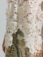 A close-up of the Social Queen Pearl Rose Guipure Lace Top, featuring white floral guipure lace with 3D rose appliqué and pearl embellishments, styled with denim jeans showcasing a faded abstract print.