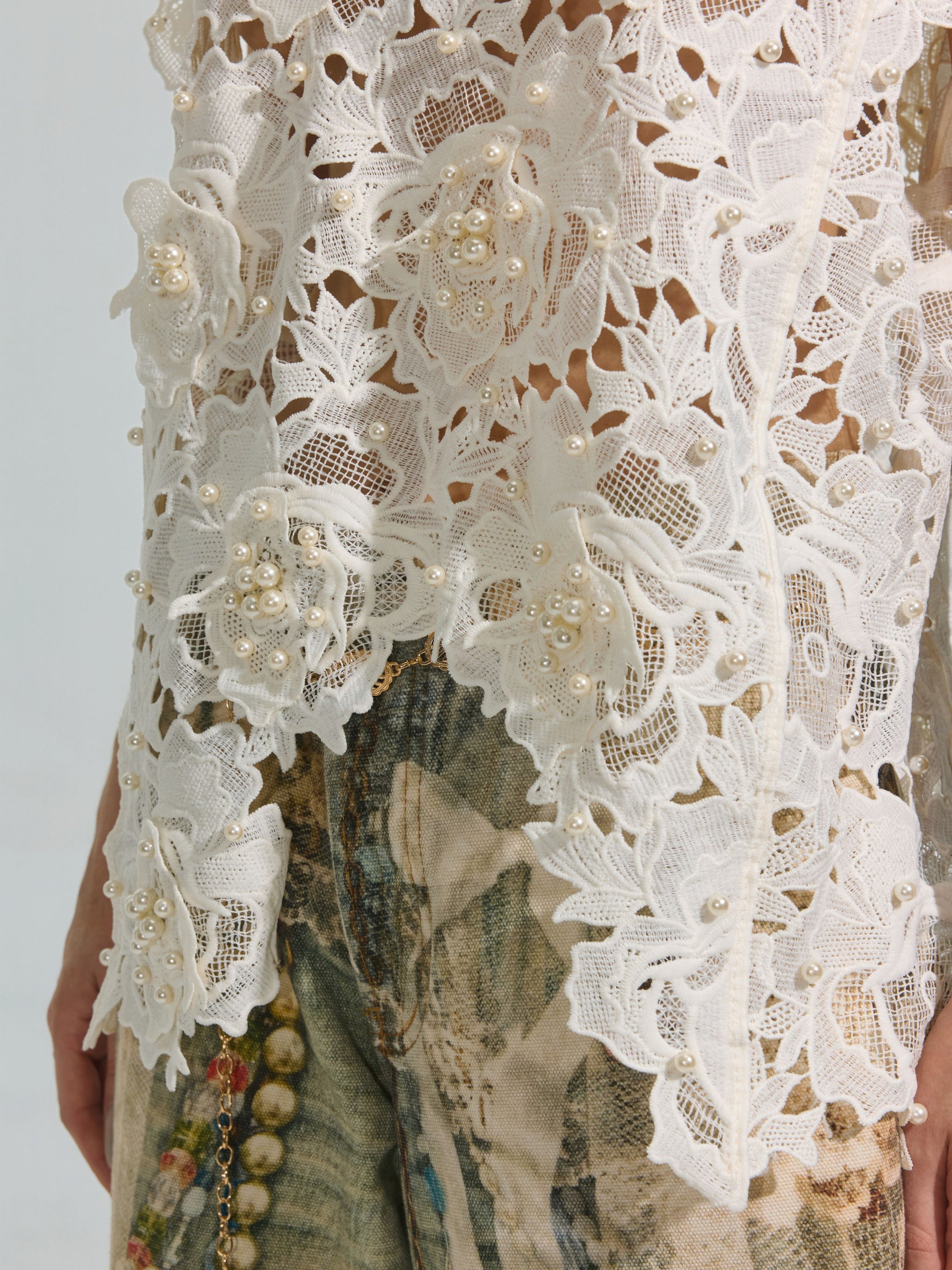 A close-up of the Social Queen Pearl Rose Guipure Lace Top, featuring white floral guipure lace with 3D rose appliqué and pearl embellishments, styled with denim jeans showcasing a faded abstract print.