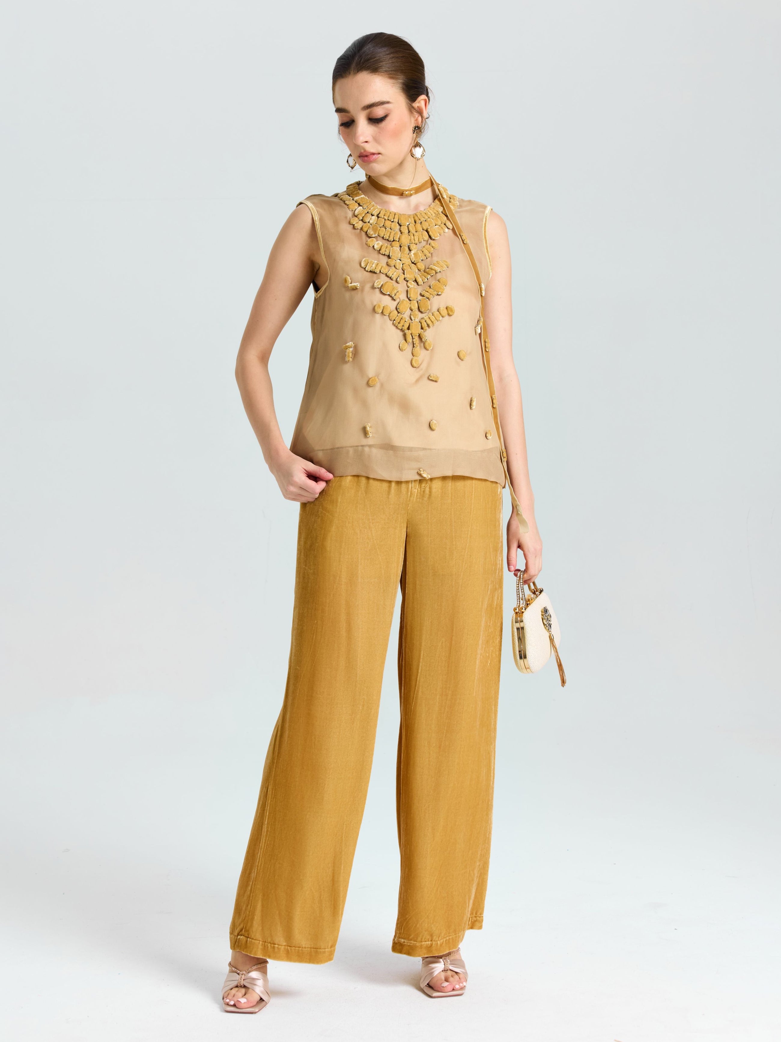 A woman stands against a plain light background wearing Social Queen's Silk Velvet Wide-Leg Pants, paired with a sleeveless beige textured top, sandals, and a small crossbody purse.