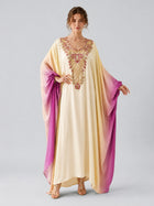 A person stands facing forward against a plain background, wearing the Social Queen Ombré Beaded Embroidered Cape Dress—its flowing cream and purple hues adorned with ornate gold and pink embroidery that echoes the elegance of a cape silhouette maxi.