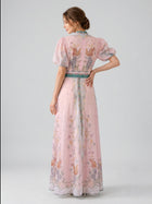 A woman stands with her back to the camera, wearing the Social Queen Peacock Botanical Print Dress, a long, sheer pink gown adorned with vibrant botanical patterns and puffed sleeves. Subtle rhinestone accents catch the light, lending an elegant touch to her ensemble. Her hair is styled in a low updo, and the look is completed with a coordinating belt.