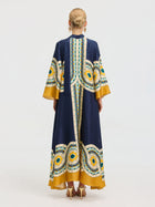 A person with blonde hair in an updo is seen from the back wearing the Social Queen Moroccan Print Silk Caftan Dress, a long robe with wide sleeves in navy blue and yellow, against a plain background.