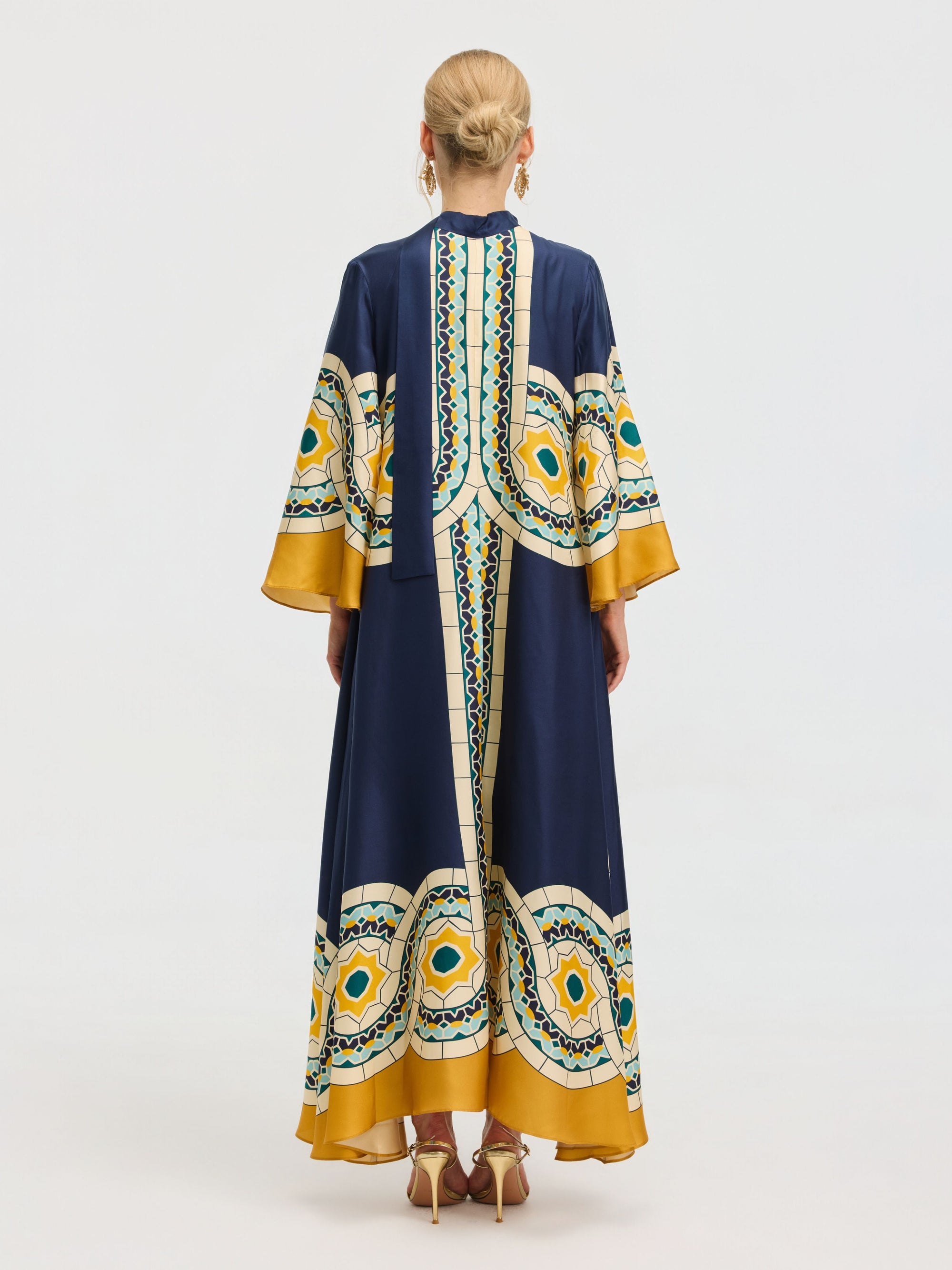 A person with blonde hair in an updo is seen from the back wearing the Social Queen Moroccan Print Silk Caftan Dress, a long robe with wide sleeves in navy blue and yellow, against a plain background.