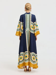 A person with blonde hair in an updo is seen from the back wearing the Social Queen Moroccan Print Silk Caftan Dress, a long robe with wide sleeves in navy blue and yellow, against a plain background.